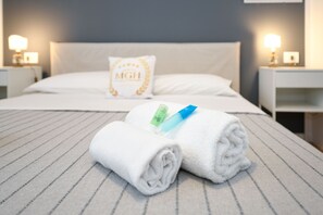 A neatly made bed with white linens and a folded towel, a pillow with 'MGH' embroidered on it, and two bedside lamps on either side of the bed. - Jardin Bardolino Apartment - Corail Apartment (Bardolino)