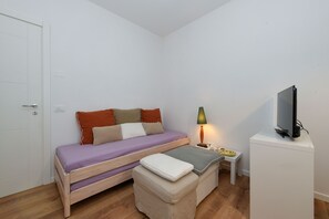 A single bed with a wooden frame, a small bedside table with a lamp, a television on a stand, and a door. - Garden Apartment Bardolino - Coral Apartment (Bardolino)