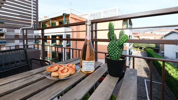 Ein Balkon mit Holztisch, einer Weinflasche, einer Auswahl an Gebäck und einem Kaktus im Topf.