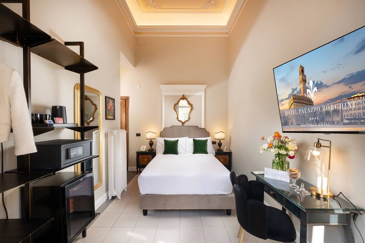 Ein Hotelzimmer mit Bett, Schreibtisch, Stuhl, Fernseher und einem Gemälde eines Gebäudes.