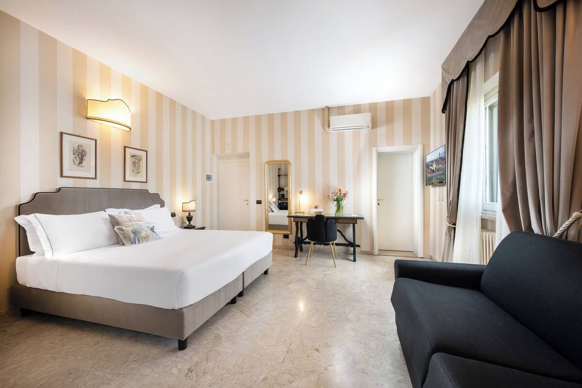 Ein Hotelzimmer mit Bett, Schreibtisch, Stuhl, einer Couch und einem großen Fenster mit Vorhängen.