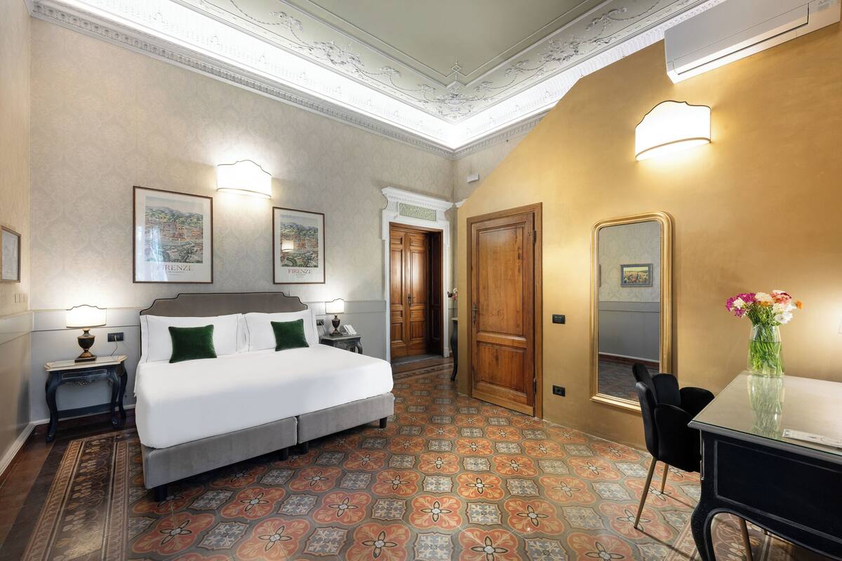 Ein Hotelzimmer mit Bett, Nachttisch, Schreibtisch, Stuhl, Spiegel und einer Vase mit Blumen.