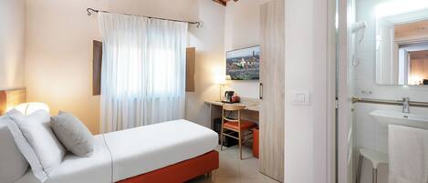 A bedroom with a bed, a desk, a chair, a TV, and a bathroom with a sink and mirror.
