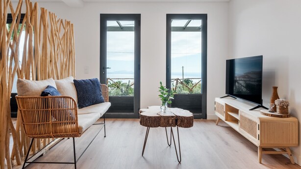Ein modernes Wohnzimmer mit einer Couch, einem Fernseher, einem runden Holztisch und Blick durch große Fenster.