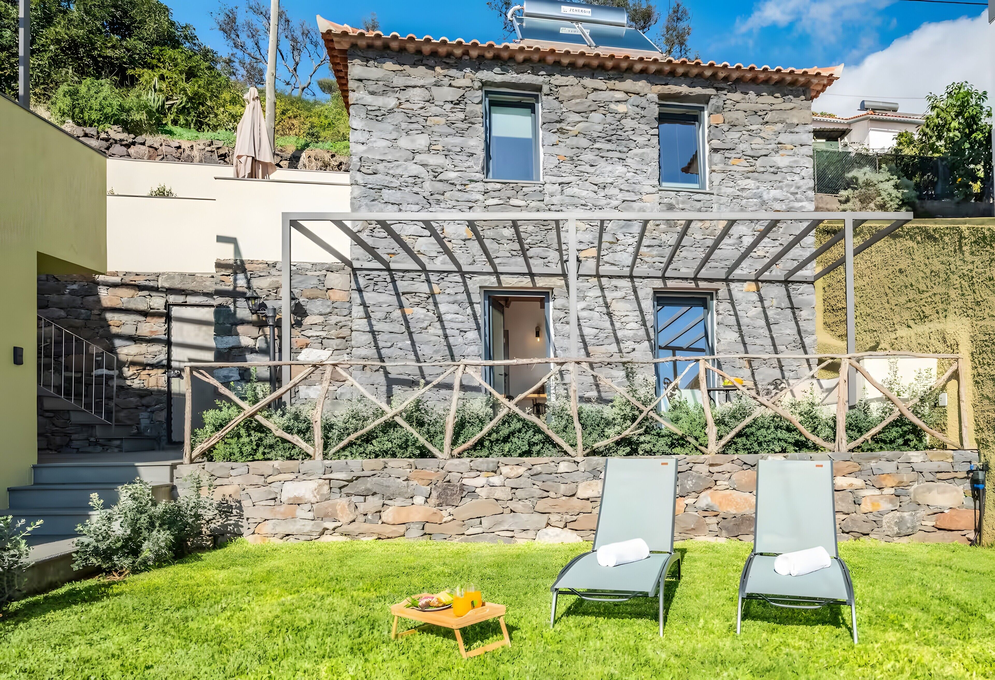A stone house with a pergola, two lounge chairs, and a small table with a drink.
