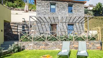Una casa de piedra con una pérgola, dos sillones de descanso y una mesa pequeña con una bebida.