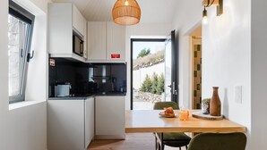A compact kitchen with white cabinets, a black countertop, a hanging light fixture, and a dining area with a wooden table and chairs. - Studio Apartment 'Ocean View Rock House  2' with Balcony and Wi-Fi (Caniço)