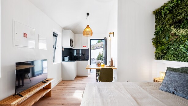 A modern room with a bed, a TV on a wooden stand, a small kitchen, and a green wall. - Studio Apartment 'Ocean View Rock House  2' with Balcony and Wi-Fi (Caniço)
