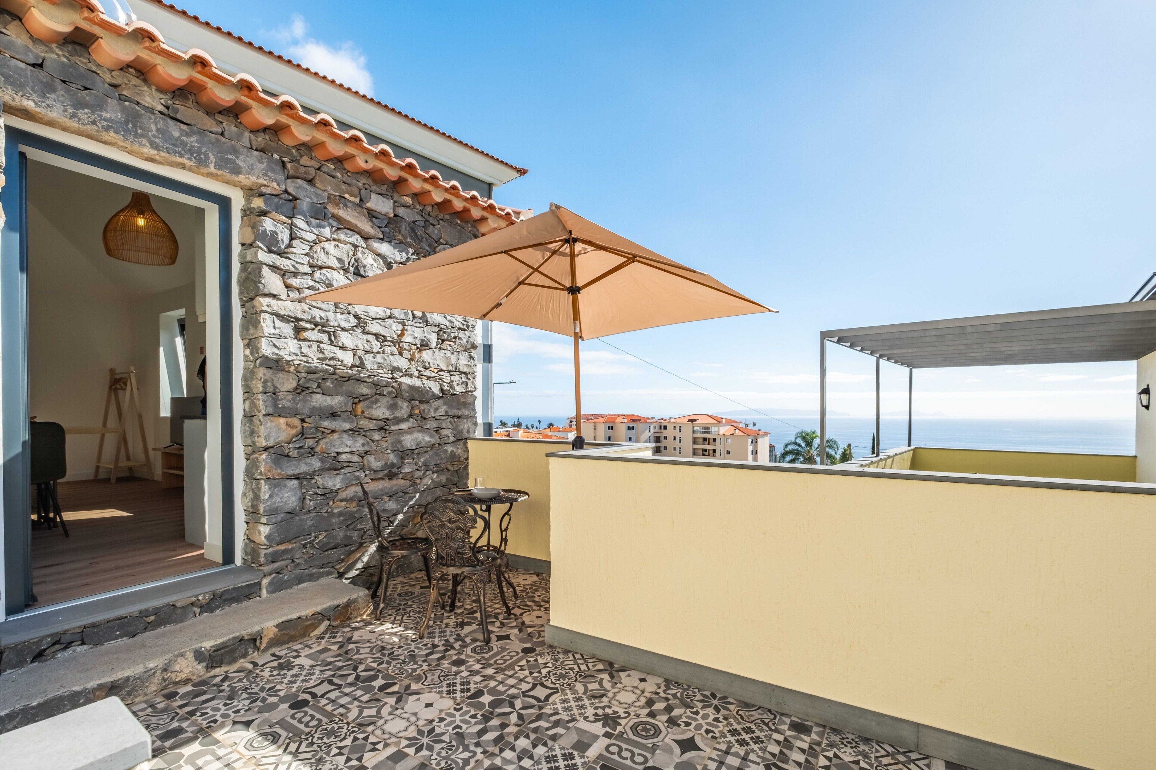 Ein Balkon mit gemustertem Boden, einer Steinsäule, einem Holztisch, einem Korbstuhl und einem großen Sonnenschirm.