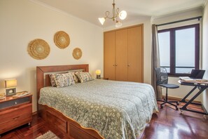 A bedroom with a large bed, wooden wardrobe, desk with chair, and a window with curtains.