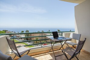 Un balcón con una computadora portátil sobre una mesa, dos sillas y vistas al mar y edificios.