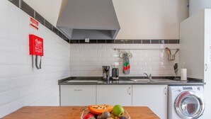 Een keuken met witte kasten, een wasmachine, een fruitschaal op het aanrecht en een brandblusser aan de muur.