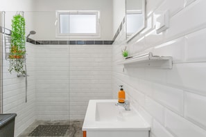 A bathroom with white tiled walls, a white sink, a window, and a shower area. - Above Funchal · A Hillside Home with Ocean Views (Funchal)