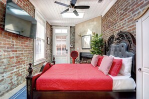 A bedroom with a brick wall, a bed with a red bedspread, a ceiling fan, and a fireplace. - Victorian Experience | Sleeps 6, Garden, 2‑Car Garage, Walk to Downtown (Savannah)