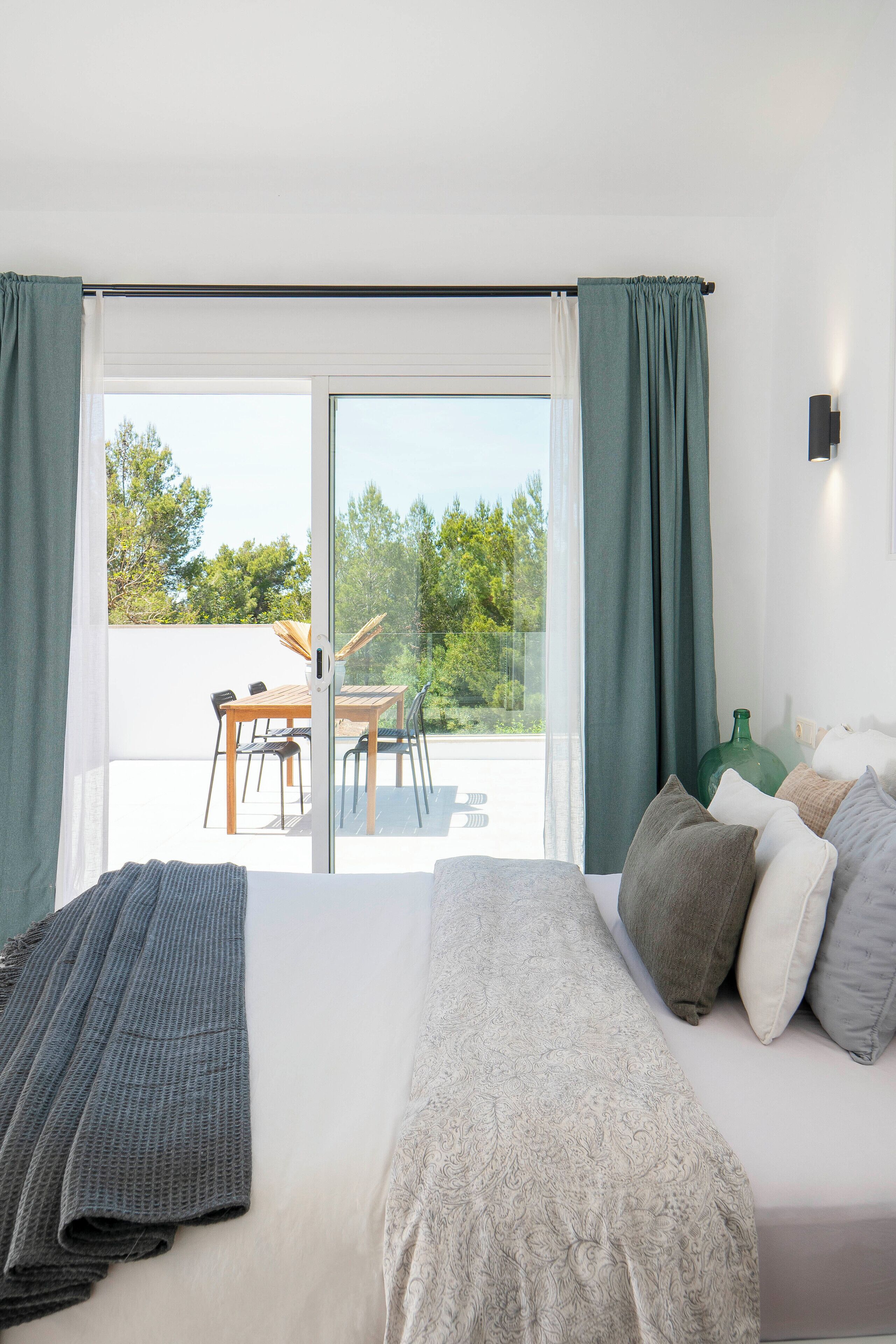 A bedroom with a bed, pillows, a blanket, and a view of an outdoor patio with a table and chairs.