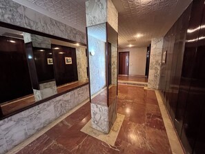 Un couloir au sol et aux murs recouverts de marbre, un comptoir d’accueil en bois et une porte au fond.