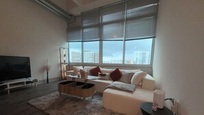 Een moderne woonkamer met een grote flatscreen-tv, een witte bank, een salontafel en een groot raam met zonwering.