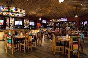 Ein Restaurant mit Holztischen und -stühlen, einer Bar mit Fernsehern und einer farbenfrohen Glasleuchte.