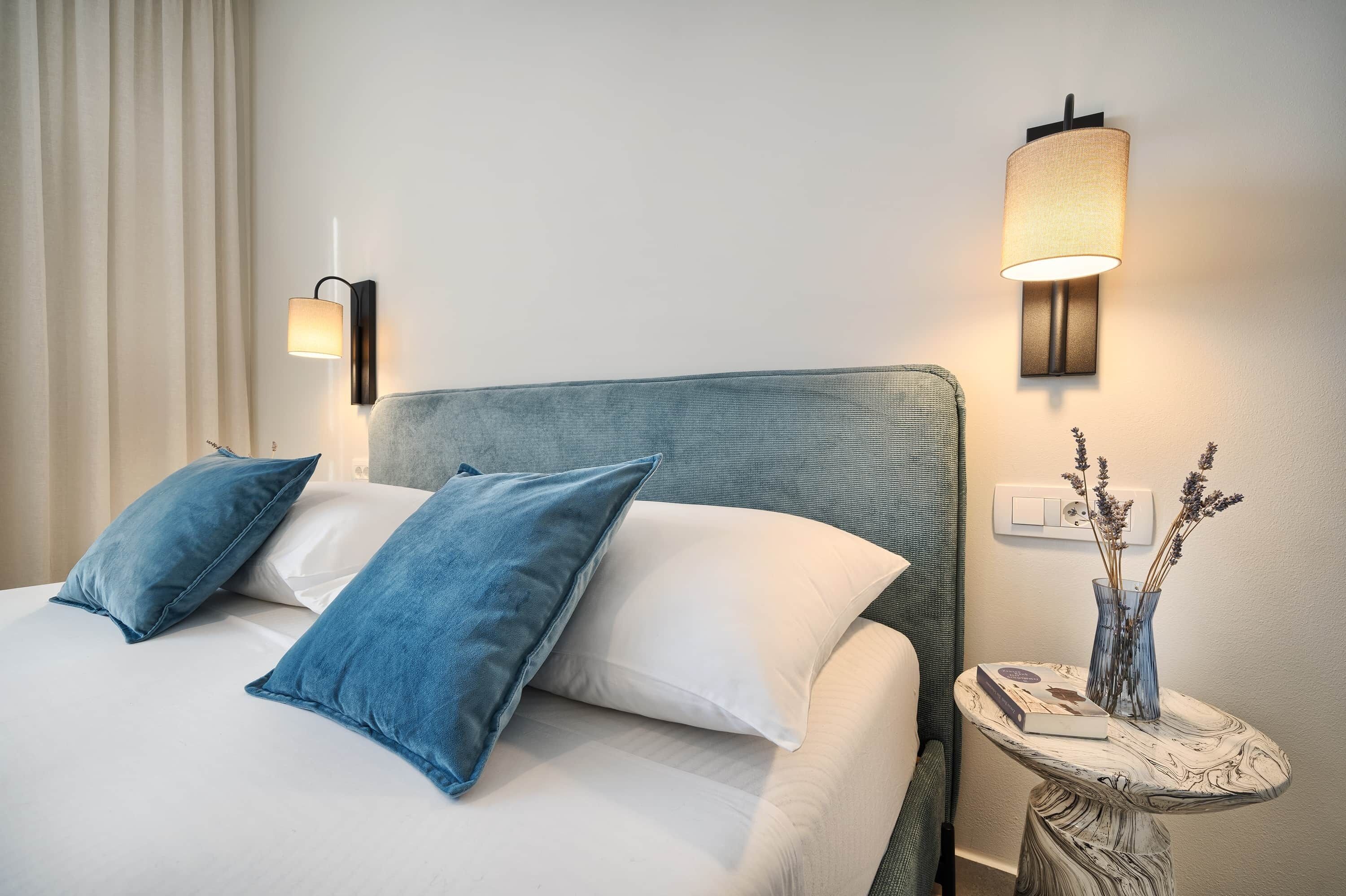 Ein ordentlich bezogenes Bett mit blauen und weißen Kissen, eine Wandlampe und ein kleiner Tisch mit Blumenvase und Buch.