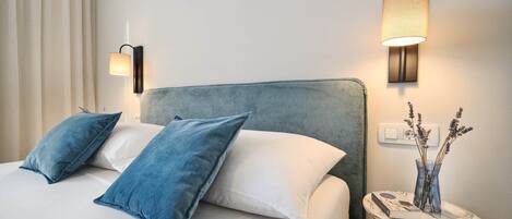 Ein ordentlich bezogenes Bett mit blauen und weißen Kissen, eine Wandlampe und ein kleiner Tisch mit Blumenvase und Buch.