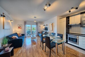 A modern living room with a sofa, dining table, and kitchen area with an oven. - Aurore's Place - Cozy Seaview Apartment (Okrug Gornji)