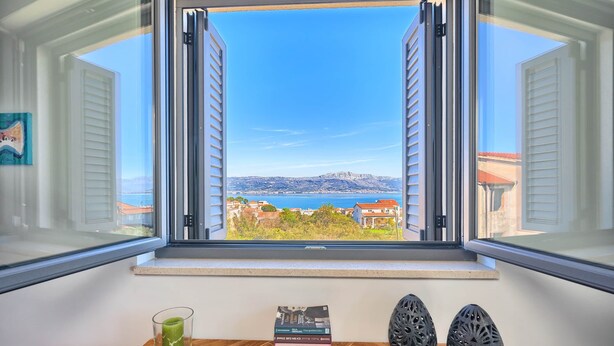 A room with a window overlooking a scenic view of a coastal area, a wooden table with books and a glass, and a red chair. - One Bedroom Villa Ming (Slatine)