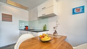 A modern kitchen with white cabinets, a glass backsplash, and a wooden dining table with a fruit bowl. - One Bedroom Villa Ming (Slatine)