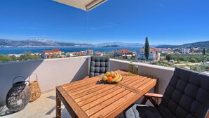 A wooden table with a fruit bowl, two wicker baskets, and a cushioned chair on a balcony overlooking a scenic view of a coastal town and mountains. - One Bedroom Villa Ming (Slatine)