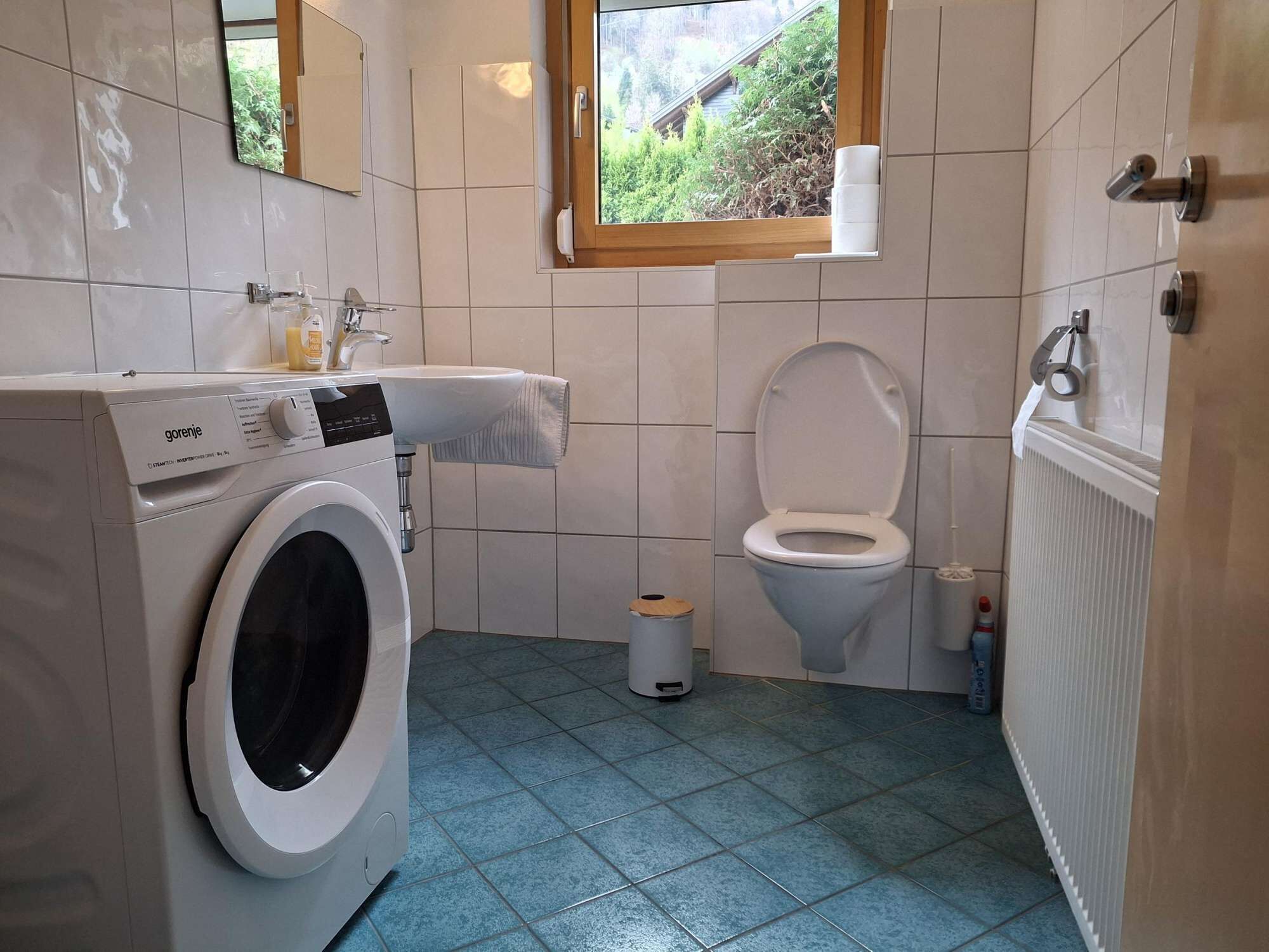 Et lille badeværelse med vaskemaskine, toilet og et vindue med udsigt til grønne omgivelser.