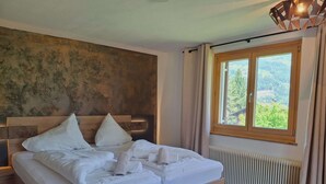 Un lit double avec du linge de lit blanc, une tête de lit en bois, une vue sur la verdure par la fenêtre et un radiateur au sol.