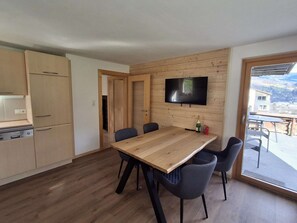 A modern kitchen with wooden cabinets, a dining table with chairs, and a TV mounted on the wall.