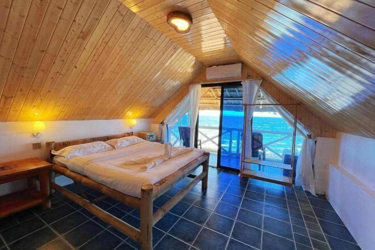 A bedroom with a wooden ceiling, a bed, a nightstand, and a view of the sea through a glass door.