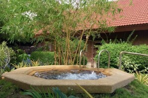 Un bain à remous en pierre, entouré de bambous et de verdure, avec une structure de toit en tuiles en arrière-plan.