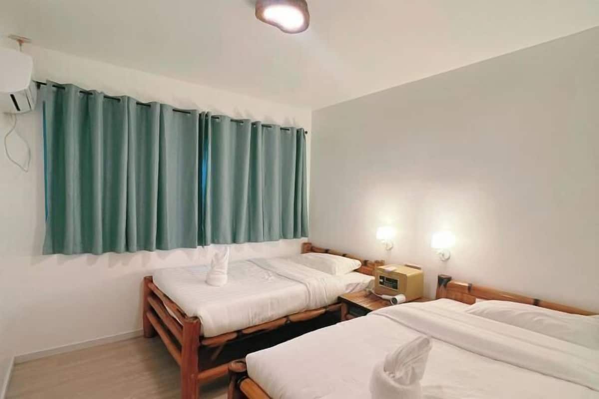 A room with two wooden beds, a window with teal curtains, and a bedside table with a lamp and a clock.