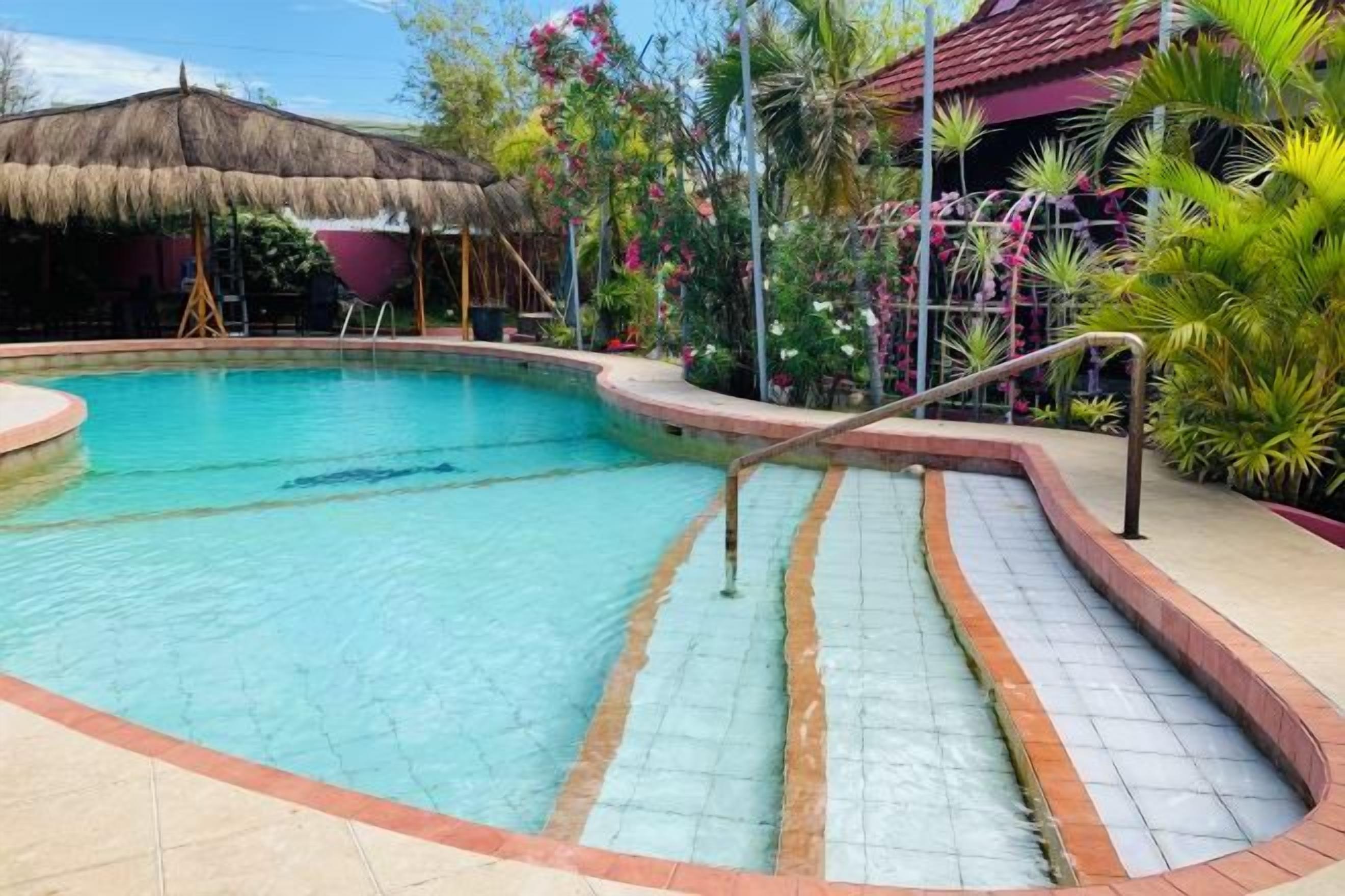 Una piscina con borde de azulejos, rodeada por un sendero de ladrillo y una estructura con techo de paja al fondo.