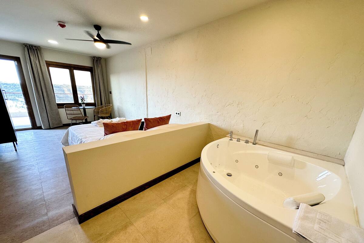 A modern hotel room with a large bed, a separate sitting area with a chair, a bathtub, and a view of the outdoors.