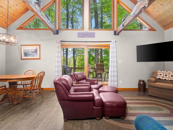 A spacious living room with a large leather sectional sofa, a dining area with wooden chairs, and a view of the forest through large windows.