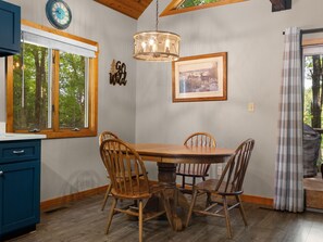 A dining area with a wooden table and chairs, a hanging light fixture, a clock, and a framed picture on the wall.