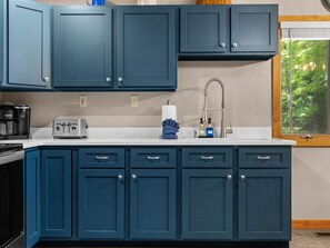 A kitchen with dark blue cabinets, a white countertop, a toaster, a coffee maker, and a window with a view of trees.