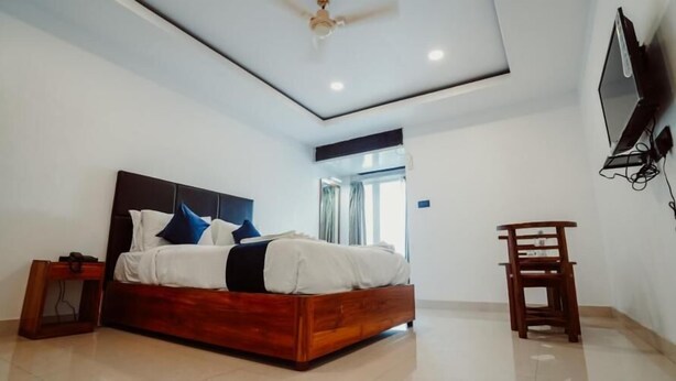A modern bedroom with a wooden bed, a ceiling fan, a TV mounted on the wall, and a small wooden chair. - Sayora Hotels (Port Blair)