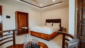 A hotel room with a wooden bed, a wooden wardrobe, a small table with glasses and a bottle, and a ceiling fan. - Sayora Hotels (Port Blair)