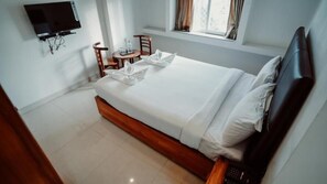 A neatly made bed with white linens, a wooden headboard, and a small table with chairs beside the window. - Sayora Hotels (Port Blair)