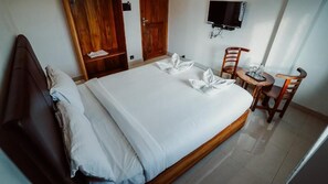 A neatly made bed with white linens and towels arranged on the bed. There is a wooden headboard, a wooden nightstand with a lamp, and a small wooden table with chairs. A television is mounted on the wall above the bed. - Sayora Hotels (Port Blair)