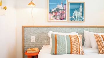 Una habitación de hotel con cama, almohadas, mesita de noche, lámpara y dos carteles enmarcados en la pared.