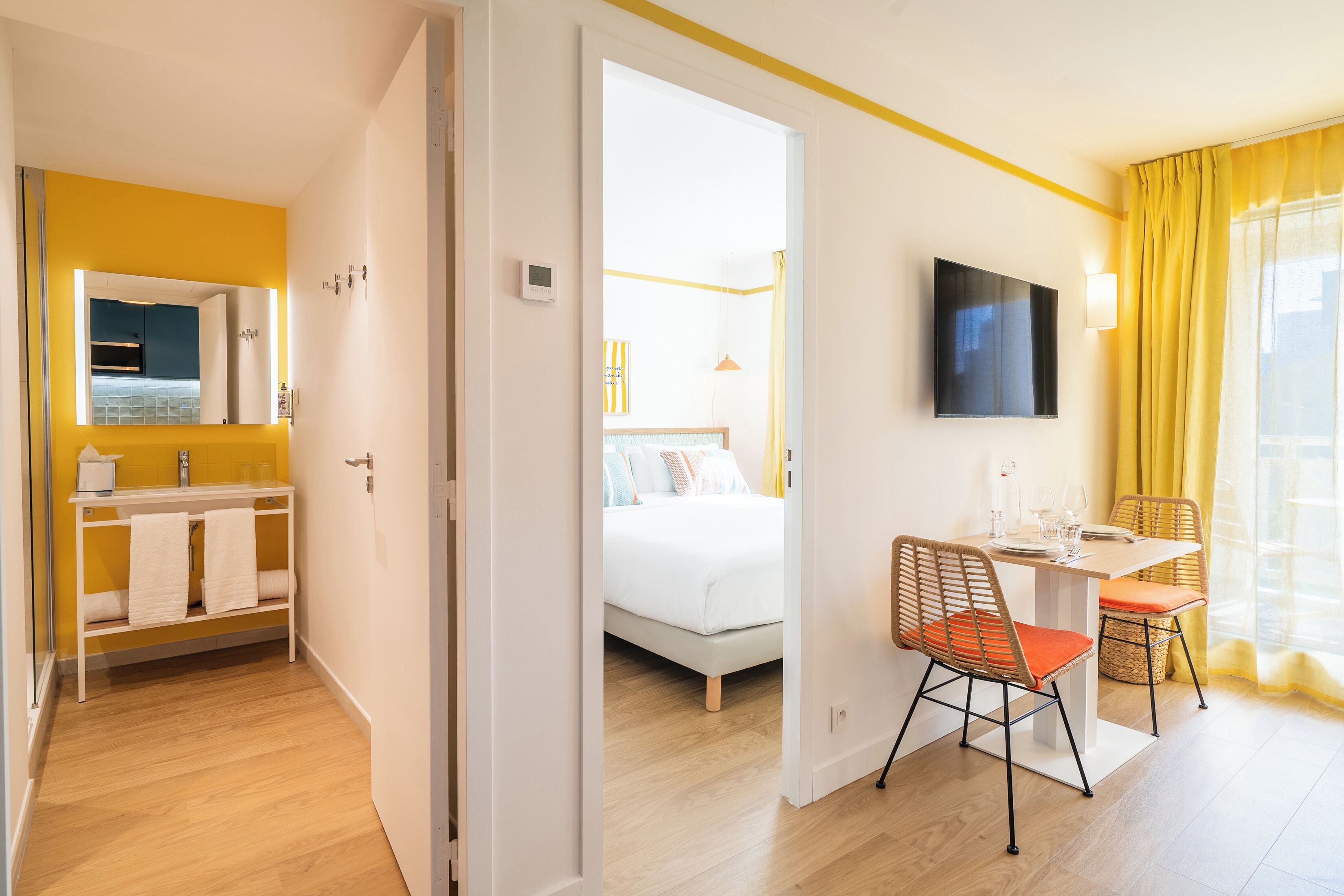 Habitación de hotel con cama, una mesa de comedor con sillas, televisor y baño con espejo y lavamanos.
