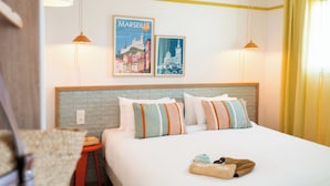 Ein Hotelzimmer mit Bett, Kissen, einem Kopfteil, einer Wand mit zwei gerahmten Bildern, einem roten Hocker und einem Fenster mit gelben Vorhängen.
