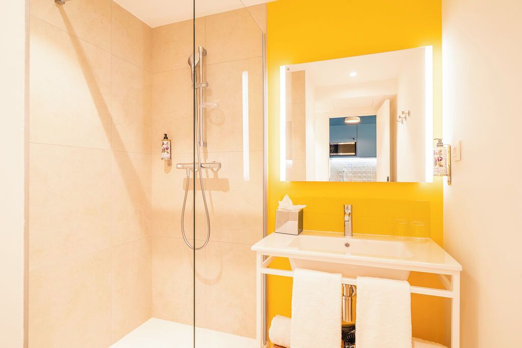 Een moderne badkamer met een douchecabine van glas, een witte wastafel en een gele accentmuur, met een spiegel die een keuken weerspiegelt.