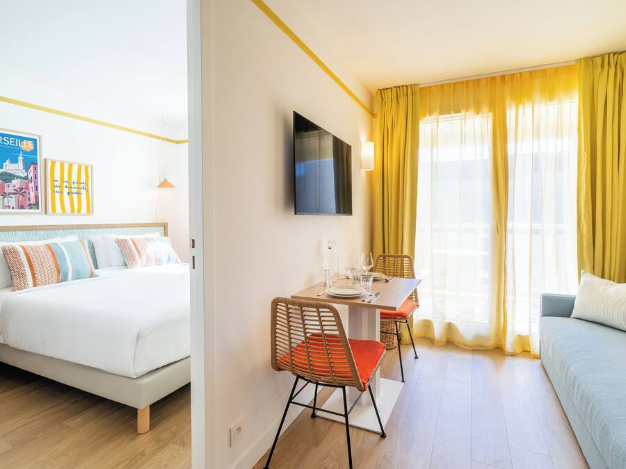 Une chambre d’hôtel comprenant un lit, un canapé, une table à manger avec des chaises, un téléviseur fixé au mur et une fenêtre avec des rideaux.