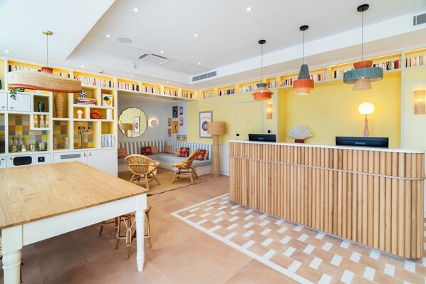 A reception area with a wooden desk, wicker chairs, a patterned rug, and a bookshelf filled with books. - Appartements Soléo Prado (Marseille)