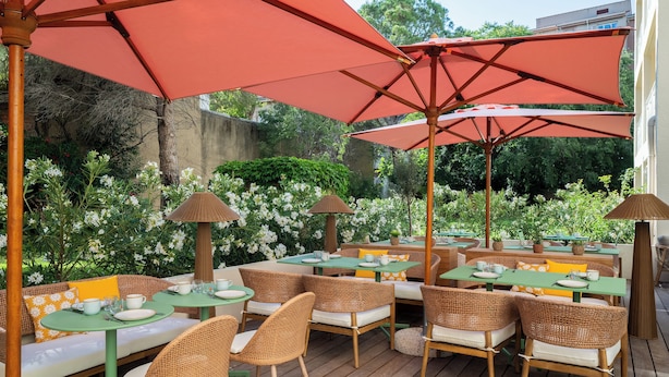 Eine Terrasse im Freien mit Korbmöbeln, grünen Tischen und roten Sonnenschirmen.